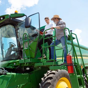 Professional farmer with a modern tractor, combine at a field in sunlight at work. Confident, bright summer colors. Agriculture, exhibition, machinery, plant production. Senior man on his machine with investor.